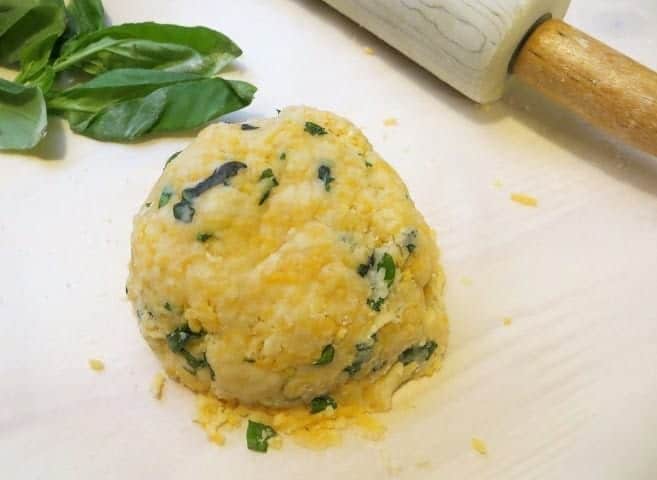 Ball of parmesan, basil, and cheddar bite dough laying on a white surface. A sprig of basil and a marble rolling pin lay in the background.