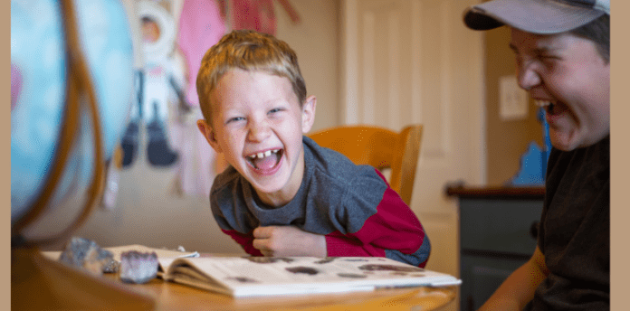 Do you need a dedicated room for homeschooling cover A laughing boy looks at the camera while leaning on a book on a table.