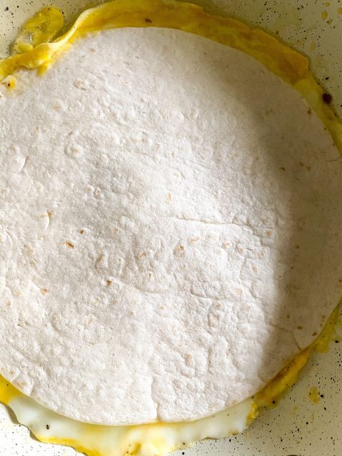 Flour tortilla laying on top of beaten egg in a frying pan