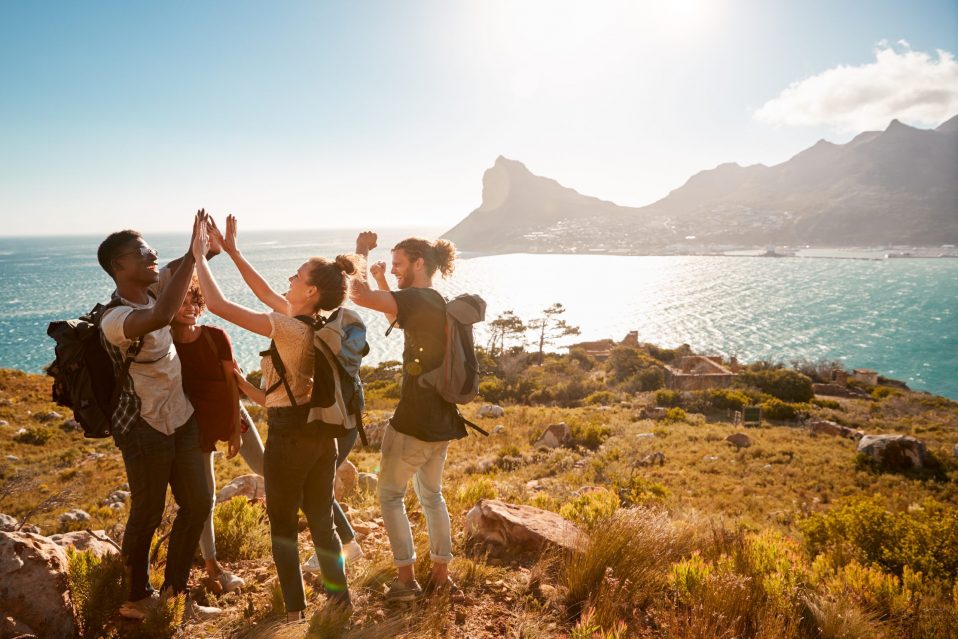 Four young people with backpacks, in front of a scenic view, high-fiving at the end of a hike