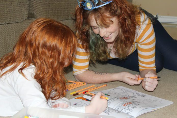 Two redheaded girls are colouring in a book on the floor. The older girl wears a tiara